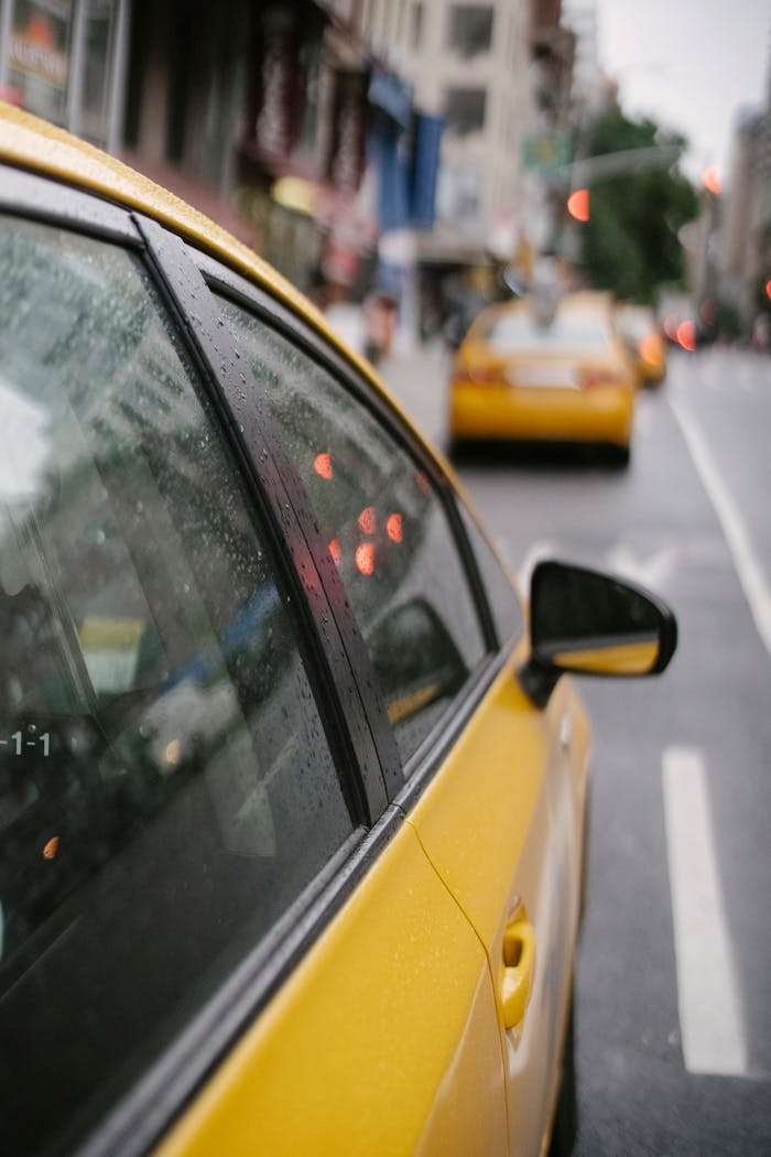 Side of yellow clean taxi with reflection in mirror driving down street with white marking on rainy day on blurred background